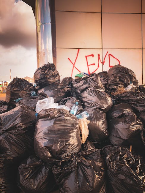 A large pile of black plastic rubbish bags, some partially torn or bulging, is stacked against a modern building with beige tiled exterior walls. The bags contain various waste items, including visibl